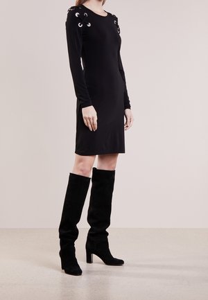 Frau trägt ein eng anliegendes schwarzes Langarmkleid mit Schnürdetails an den Schultern und schwarze kniehohe Stiefel mit Absatz, steht auf einem schlichten Boden.