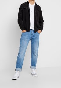 Camisa negra abotonada sobre una camiseta blanca, jeans azul claro con puños enrollados y zapatillas blancas de caña alta.