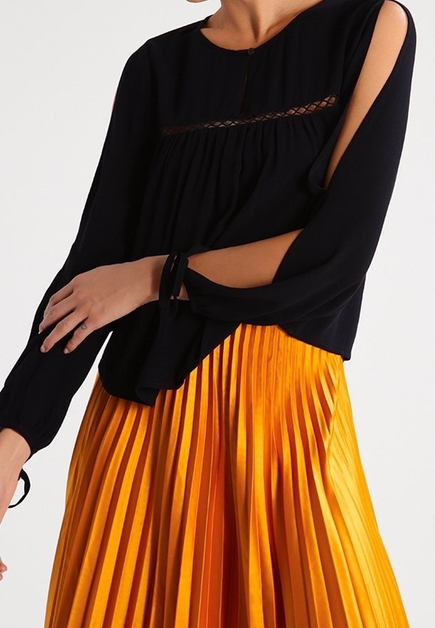Blusa nera con maniche lunghe caratterizzata da aperture e un dettaglio in pizzo orizzontale, abbinata a una gonna arancione plissettata, creando un look a strati.