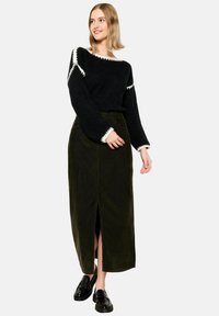 Čierny pletený sveter s bielym lemovaním, kombinovaný s tmavozelenou kordurovouv maxi sukňou s predným rozparkom. Model nosí čierne topánky.