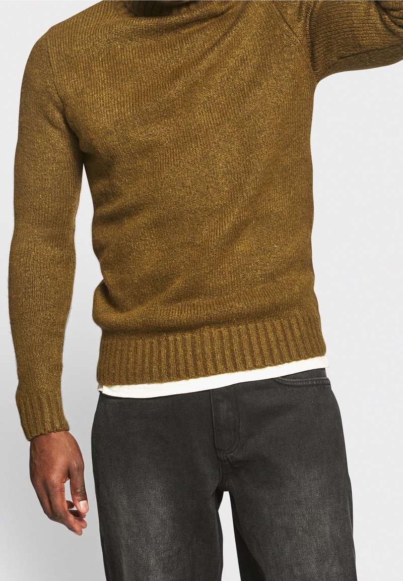 Homme portant un pull en maille côtelée vert olive sur une chemise blanche avec un jean noir, sur un fond clair uni.