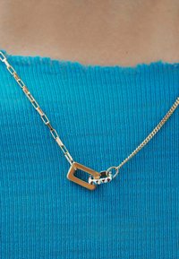 Collar de cadena de oro con un cierre rectangular, hecho de metal enlazado, descansando sobre un fondo de tela turquesa texturizada.