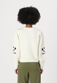 Maglione lavorato a maglia di colore crema con maniche a sbuffo, presenta ricami floreali neri sulle maniche e un orlo a coste. Morbido al tatto, vestibilità rilassata.