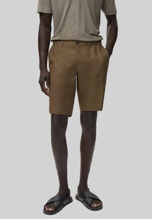Mand iført brune shorts, lysebrun kortærmet skjorte og sorte sandaler med krydsede stropper står mod en ensfarvet baggrund.