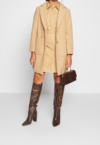 Abrigo de lana beige con dos bolsillos frontales sobre un vestido marrón claro con detalles de ojales, combinado con botas altas de serpiente marrón y un bolso pequeño.