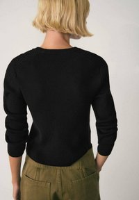 Pull noir en maille avec texture côtelée, manches ajustées et encolure arrondie, vu de dos, associé à un pantalon vert olive.