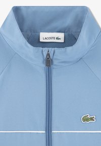 Ανοιχτό μπλε μπουφάν με φερμουάρ, διαθέτει γιακά από δίχτυ, λευκές ρίγες ως διακοσμητικά στοιχεία και πράσινο κεντημένο λογότυπο στο στήθος. Ανθεκτική υφή υφάσματος.