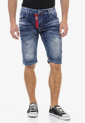 Mann trägt mittel-lange ausgewaschene Jeansshorts mit rotem Reißverschluss-Detail, schwarzes T-Shirt und schwarze Low-Top-Sneaker mit weißen Schnürsenkeln.