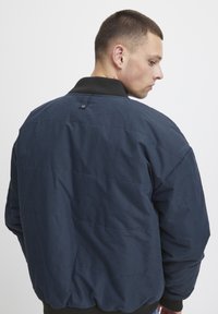 Tamsiai mėlyna izoliacinė bomber tipo striukė su tekstūruotu audiniu, juodu rainu apykakle ir mygtuko detale gale. Matomas siuvinėjimo raštas.