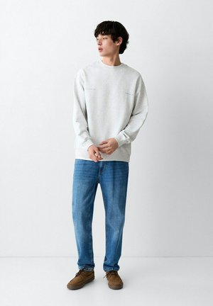 Giovane uomo in piedi, indossa una felpa grigio chiaro, jeans blu e scarpe marroni, guarda verso sinistra con uno sfondo bianco semplice.