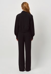 Zwarte jas met een elastische taille, lange mouwen en kantaccenten. Gecombineerd met zwarte wijde broek. Soepele stofstructuur.