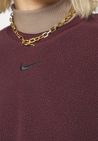 Bordeauxfärgad fleece-tröja med svart Nike-logga, ribbad beige polotröja under, och guldhalsband. Strukturerad tygdetalj synlig.