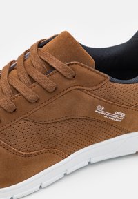Brown suede sneaker with perforated details, flat brown laces, padded collar, and white rubber sole. White label with text on the side.