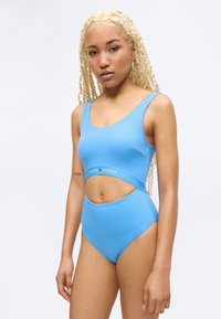 Νεαρή γυναίκα με μακριές ξανθές πλεξούδες που φοράει γαλάζιο ολόσωμο μαγιό με κοψίματα και το λογότυπο Tommy Hilfiger, στέκεται μπροστά σε ένα απλό λευκό φόντο.