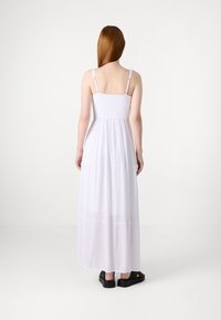 Vit maxiklänning med justerbara band, smockad bodice och flödande kjol, tillverkad av lättviktig tyg; bärs med svarta sandaler.