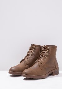 Brown ankle boots made of faux leather, featuring metal eyelets, laces, a side zipper, and a low stacked heel with a textured sole.