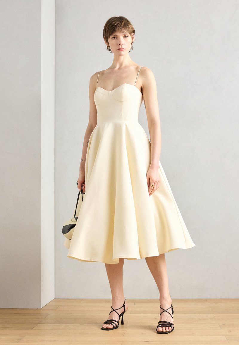 Cremfarbene Satin-Langarmkleid mit tailliertem Oberteil, ausgestelltem Rock, verstellbaren dünnen Trägern und glatter Textur. Kombiniert mit schwarzen Riemchen-High Heels.
