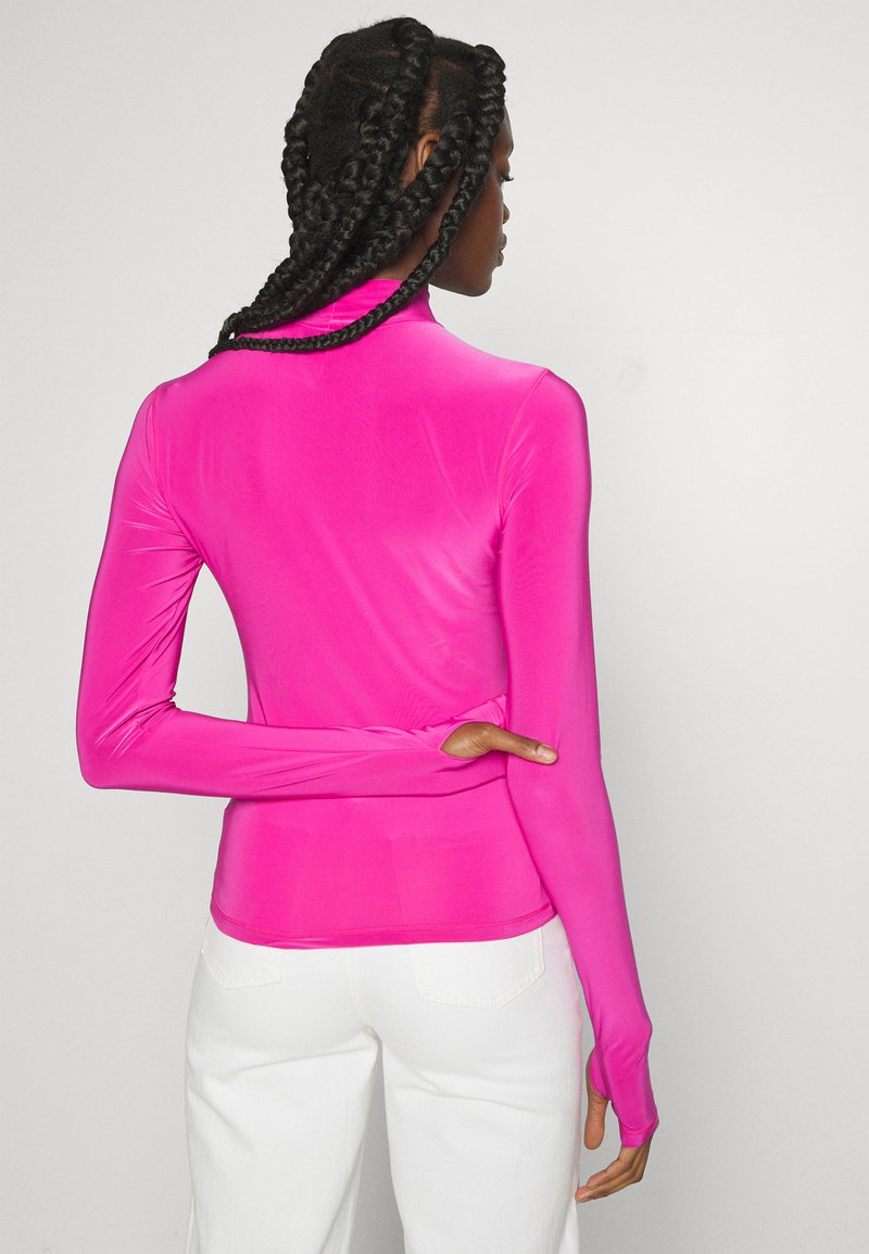 Top ajusté à manches longues de couleur rose vibrante, en matériau lisse et extensible. Il présente un col haut et des trous pour les pouces pour plus de couverture des manches.
