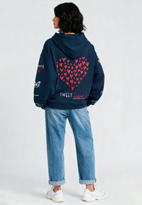 Marineblauwe hoodie met een rood hartpatroon, de tekst "SWEET heart" en vlinderafbeeldingen; gecombineerd met lichtblauwe denim en witte sneakers.