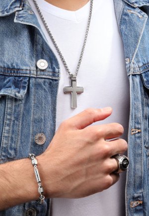 Silver cross pendant necklace hangs from a metallic chain; paired with a silver bracelet and a black-accented ring, worn over a white shirt.