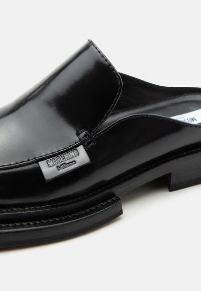 Μαύρο γυαλιστερό δερμάτινο μοκασίνι χωρίς κορδόνια με χαμηλό τακούνι, με ασημένια πλακέτα λογότυπου Moschino Milano στο πλάι.