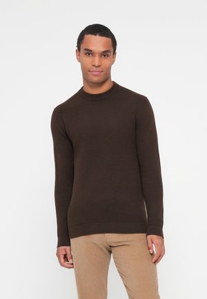 Bruine gebreide trui met een ronde halslijn en geribde manchetten. Strakke pasvorm met een gladde textuur. Gedragen met lichtbeige broek.