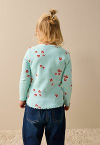 Vaaleansininen ribbineulottu pitkähihainen paita, jossa on aallotettu helma ja pieniä punaisia mansikkakuvioita. Yhdistetty tummiin denim-housuihin.