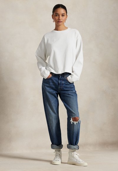 Ung kvinde stående med hænderne i lommerne, iført en hvid langærmet sweatshirt, revnede blå jeans og hvide høje sneakers.