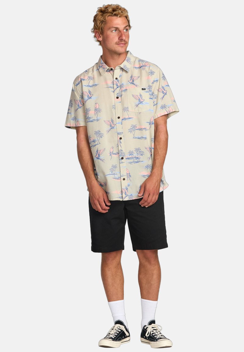 Camisa de corte corto, beige, con botones y un estampado tropical que presenta pájaros rosas y azules y palmeras. Combinada con pantalones cortos negros y zapatillas.