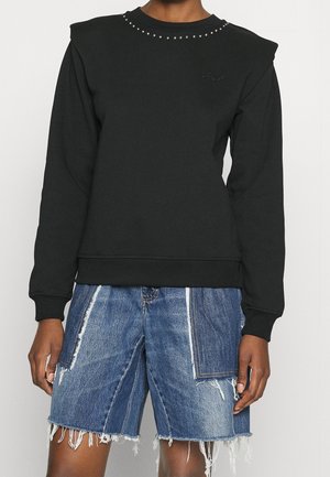 Black long-sleeve sweatshirt with silver studs on collar, paired with frayed, patchwork denim shorts on a standing person.