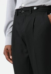 Zwarte broek met een getailleerde pasvorm, voorzien van twee plooien aan de voorkant, riemlussen en een zilveren knoopsluiting. Gemaakt van een gladde stof.