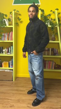 Μαύρο μπουφάν με φερμουάρ και υφή, ανοιχτό μπλε τζιν και μαύρα παπούτσια. Το φόντο έχει κίτρινους τοίχους και πράσινα ράφια με φυτά.