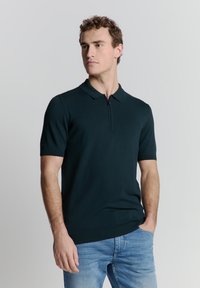 Donkergroene polo met korte mouwen en een rits aan de voorkant, gedragen met lichtblauwe jeans; gladde textuur en slim-fit ontwerp.
