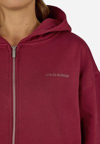 Corta-vento burdeos com fecho éclair, feito de um tecido suave; possui um grande capuz, fecho frontal e o logótipo "PEGADOR" impresso no peito.