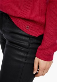 Pull rouge côtelé avec un accent rond argenté, porté sur un jean noir, montrant un gros plan sur la manche et le détail latéral.