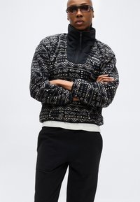 Persoană îmbrăcată cu un polar cu fermoar, negru și bej, cu pantaloni negri și ochelari de soare, cu brațele încrucișate, pe un fundal alb simplu.