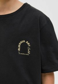 Camiseta de algodón negra con mangas cortas, que presenta un gráfico impreso en dorado de "ALMERIA ART Exhlb." cerca de la zona del pecho, con cuello redondo.