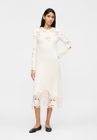 Femeie purtând o rochie midi albă cu mâneci lungi și detalii complicate din dantelă la mâneci, decolteu și tiv, asortată cu pantofi negri comozi, stând în picioare.