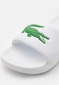 Tongs blanches avec une semelle texturée, arborant un logo en caoutchouc crocodile vert sur la sangle. Matériau lisse avec un design large.