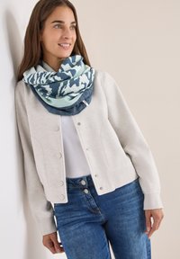 Lysegrå cardigan med knapper, hvid top og blå mønstret halsedisse; modellen poserer mod en neutral baggrund. Denim jeans er synlige.