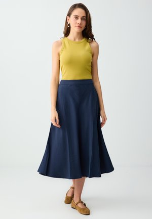 Kobieta ubrana w żółtą bluzkę bez rękawów, granatową spódnicę midi oraz brązowe płaskie buty, stoi na tle jednolitego jasnego tła.