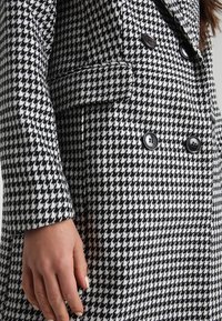 Schwarze und weiße Hahnentrittmuster-Jacke mit doppelreihigem Design, schwarzen Knöpfen und spitzem Kragen. Der Stoff wirkt strukturiert.