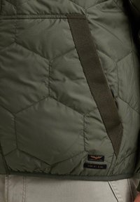 Grüner Steppjacke mit sechseckigem Muster, mit strukturierten Oberflächen, kontrastierenden Trägern und einem kleinen Etikett nahe dem Saum.