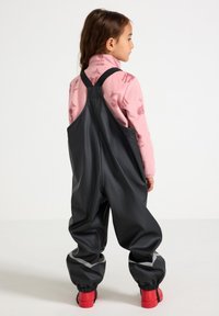 Sorte vandtætte overalls med reflekterende detaljer, elastiske manchetter og justerbare stropper, parret med en pink mønstret langærmet bluse og røde støvler.