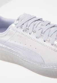 Gros plan d'une basket blanche et argentée avec une tige texturée, des lacets blancs et une semelle en caoutchouc transparente sur fond blanc.
