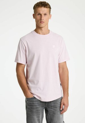 Jonge man staand met handen in zijn zakken, draagt een lichtroze T-shirt met korte mouwen en vervaagde grijze jeans tegen een witte achtergrond.