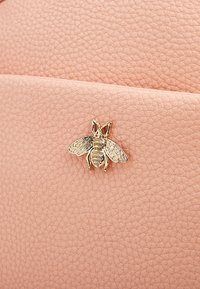 Pelle rosa chiaro testurizzata con un emblema d'oro a forma di ape. Il design presenta cuciture sottili e una finitura liscia e resistente.