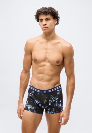 Jeune homme torse nu aux cheveux bouclés, portant un boxer noir avec un motif floral bleu, sur un fond clair uni.