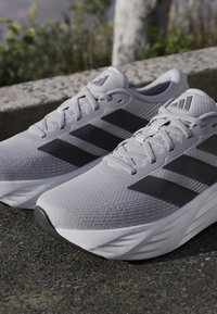 Ζευγάρι γκρι αθλητικά παπούτσια Adidas με μαύρες ρίγες τοποθετημένα έξω σε άσφαλτο κοντά σε πέτρινο κράσπεδο και πράσινο.