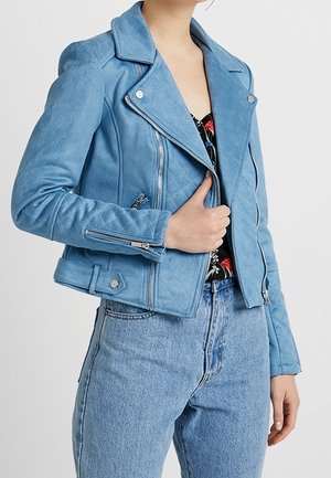 Femme portant une veste motard en daim bleu clair avec des manches matelassées, des fermetures éclair argentées et un jean bleu clair taille haute, tenant le revers de la veste.